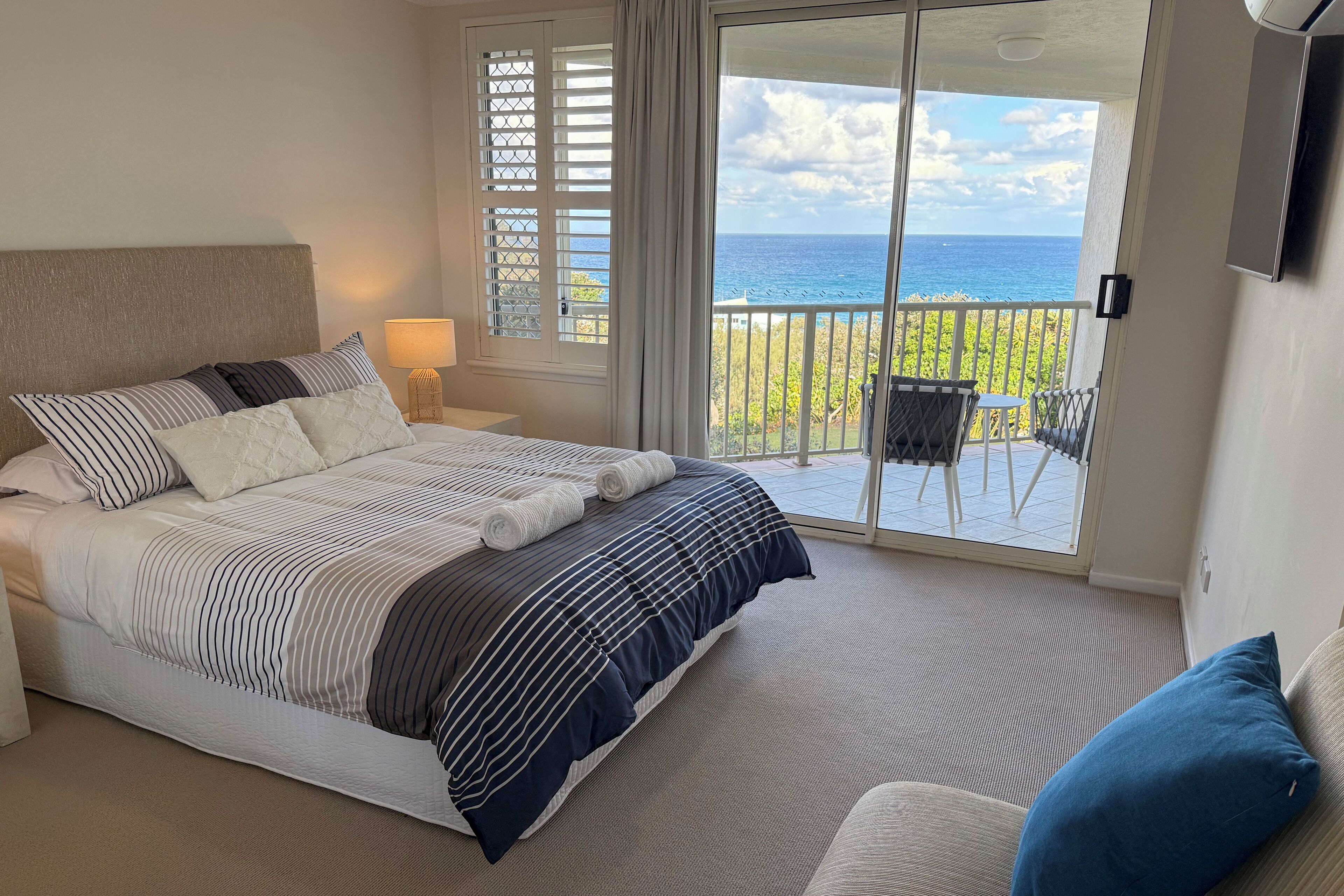 A bedroom with a bed, a nightstand, a lamp, a chair, and a balcony with a view of the ocean.