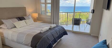 A bedroom with a bed, a nightstand, a lamp, a chair, and a balcony with a view of the ocean.