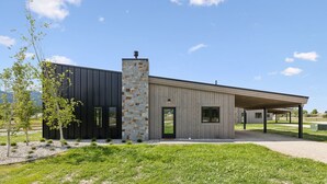 Un edificio moderno con chimenea de piedra, fachada oscura y extensión de madera con porche cubierto.