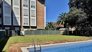 Una piscina con borde de azulejos, césped y un edificio de fachada de ladrillo con múltiples ventanas.