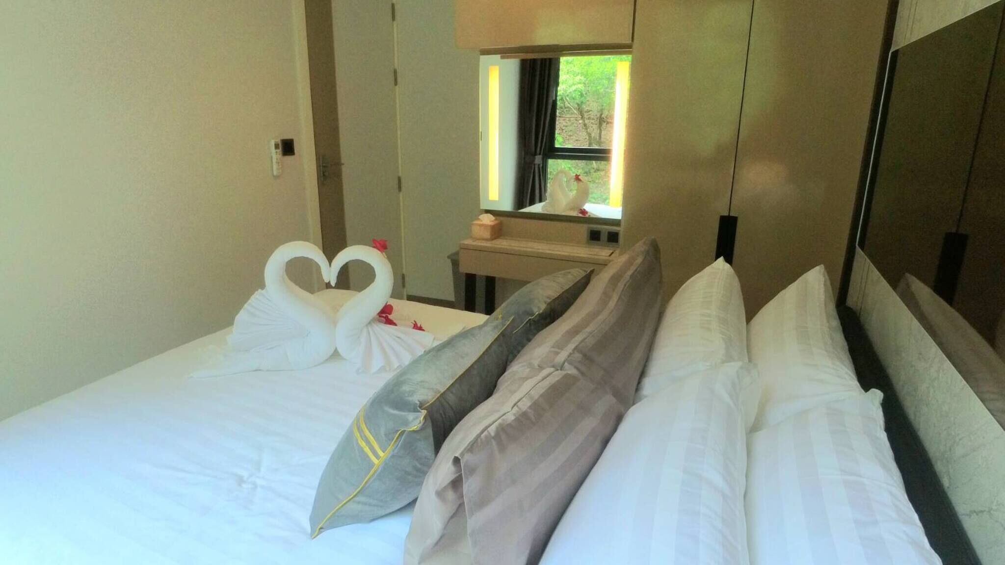 Habitación de hotel con cama, almohadas, mesita de noche, espejo y ventana con vista a la vegetación.