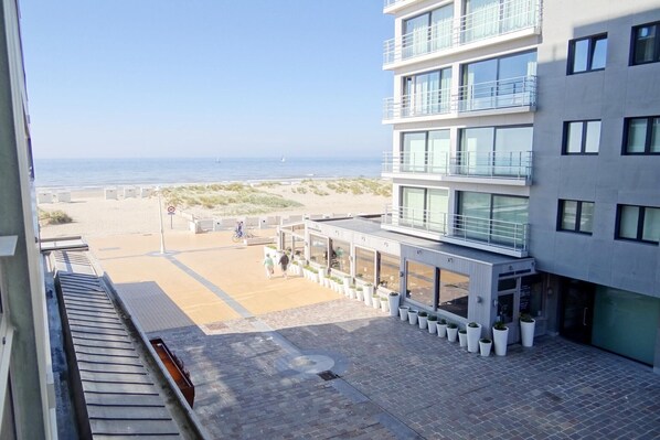 A modern building with multiple floors, large windows, and a balcony. In front of the building, there is a paved area with potted plants and a pathway leading to a beach. - Apartment in Nieuwpoort near the Beach (Nieuwpoort)
