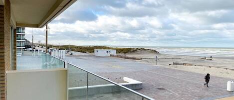 Strandudsigt fra en balkon med gelænder i glas, en sandstrand og en overskyet himmel.