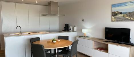 Eine moderne Küche mit weißen Schränken, einer zentralen Kochinsel mit Sitzgelegenheiten, einem eingebauten Herd und einem Flachbildfernseher auf einer Holzkonsole.