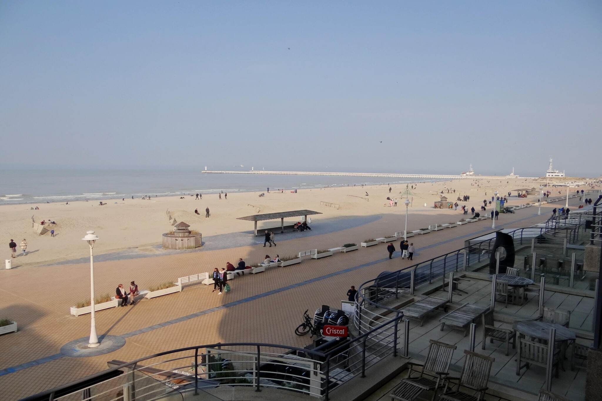 Un paseo marítimo frente a la playa, con vista despejada al mar, un muelle a lo lejos y una zona de descanso cubierta.