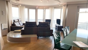 A modern living room with a curved sofa, a glass dining table, and a view of the city through large windows.