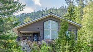 A modern cabin with a large window, surrounded by dense forest and rocky terrain.
