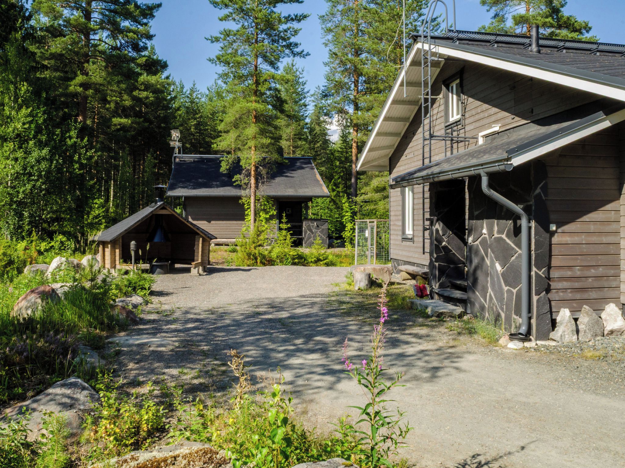 A cabin with a stone exterior, a wooden porch, and a gravel driveway surrounded by trees.