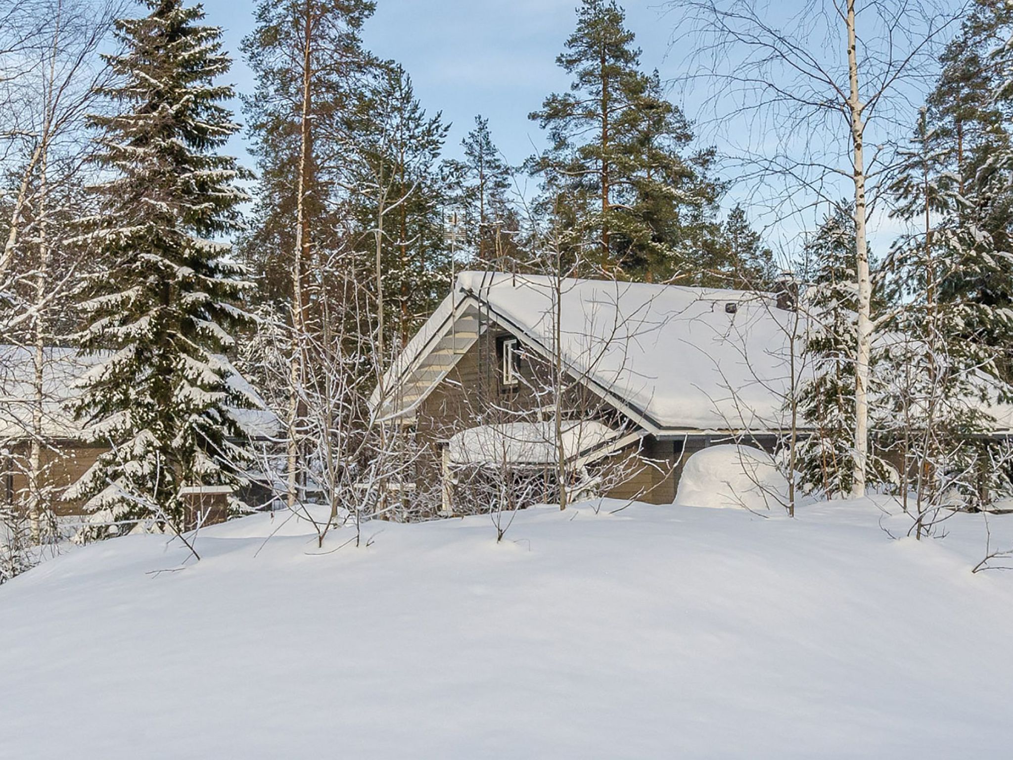 Una cabaña de techo empinado, rodeada de árboles cubiertos de nieve y un cielo despejado.