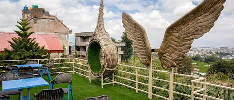Una terraza en la azotea con mobiliario de mimbre, una estructura espiral verde singular y grandes esculturas aladas.