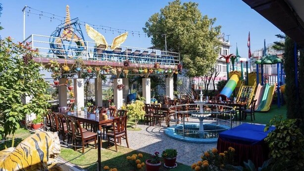 An outdoor dining area with tables and chairs, a playground with slides, and a decorative fountain. - Paradise Hills Resort Hotel (Thaiba)