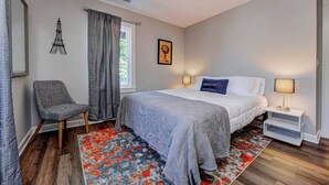 A bedroom with a bed, a chair, a nightstand, a lamp, and a window with curtains. - Chic Spacious Downtown Retreat (Indianapolis)