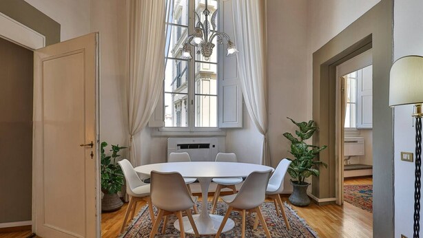 A dining area with a round table, white chairs, a chandelier, and a view of buildings through a window with curtains. - Uffizi View (Firenze)