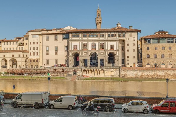 A historic building with a prominent tower, a river in the foreground, and a parking area with various vehicles. - Uffizi View (Firenze)
