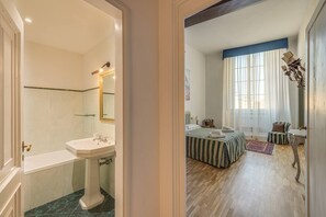 A bathroom with a pedestal sink and a mirror, a bedroom with a bed, a chair, and a window with curtains. - Uffizi View (Firenze)