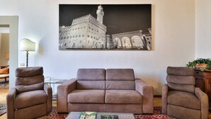 A living room with a sofa, two armchairs, a coffee table with books, and a large black and white cityscape painting on the wall. - Uffizi View (Firenze)