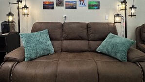 Eine braune Ledersofa mit zwei türkisfarbenen Kissen, neben zwei schwarzen Stehlampen mit klaren Gläsern, vor einer weißen Wand mit fünf gerahmten Landschaftsbildern.