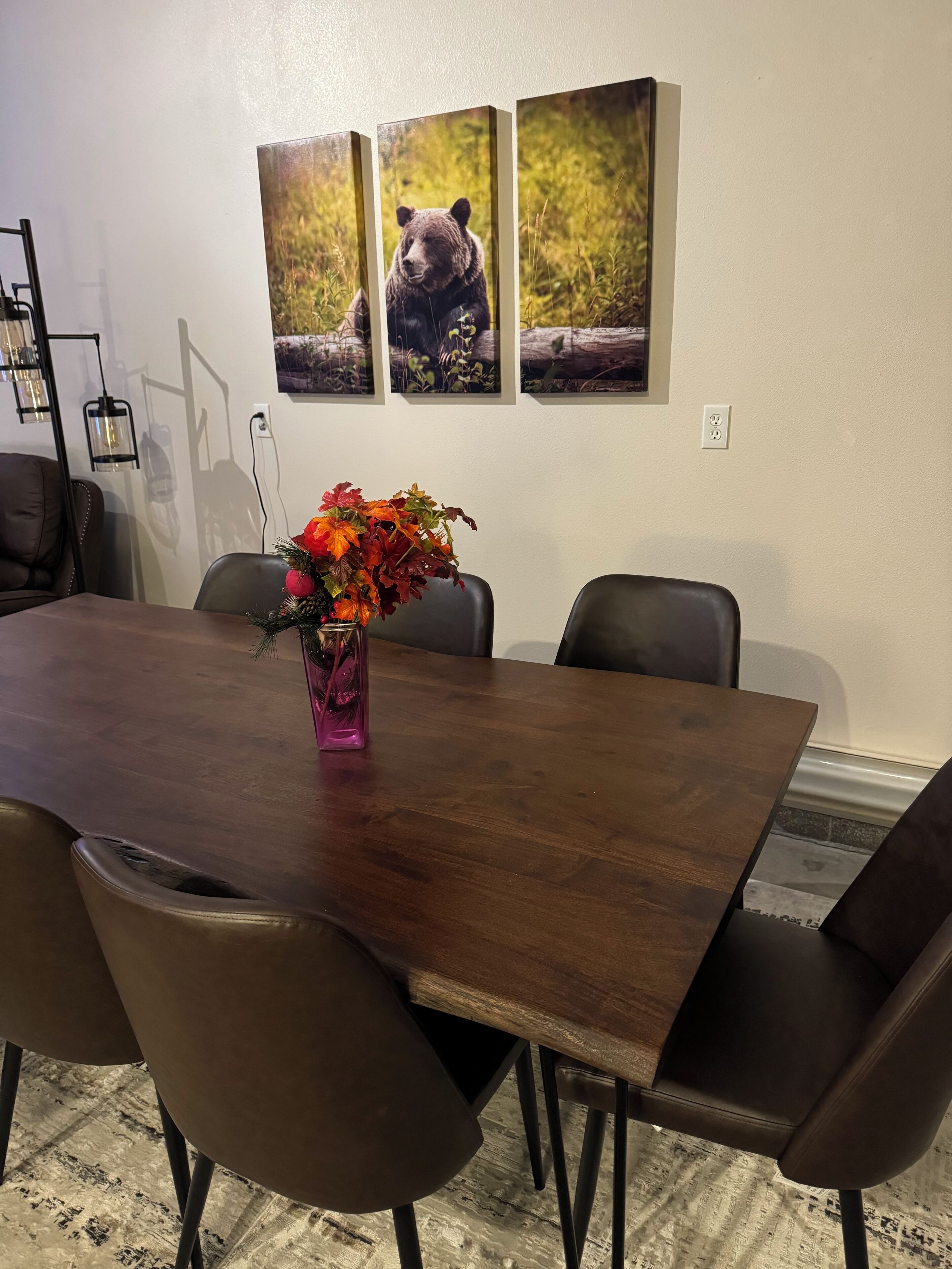 Un coin repas avec une table en bois, six chaises et un vase de fleurs. Trois tableaux encadrés représentant des ours sont accrochés au mur.