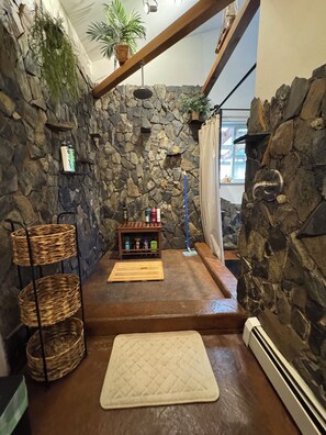 Un baño con pared de piedra, un banco de madera, un estante pequeño con artículos de aseo y una alfombra blanca para el baño.