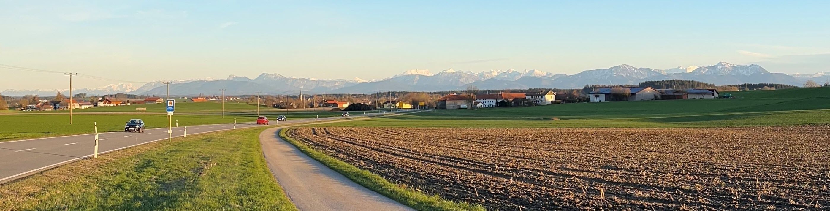 A rural road with a field and houses in the background, and a mountain range in the distance.