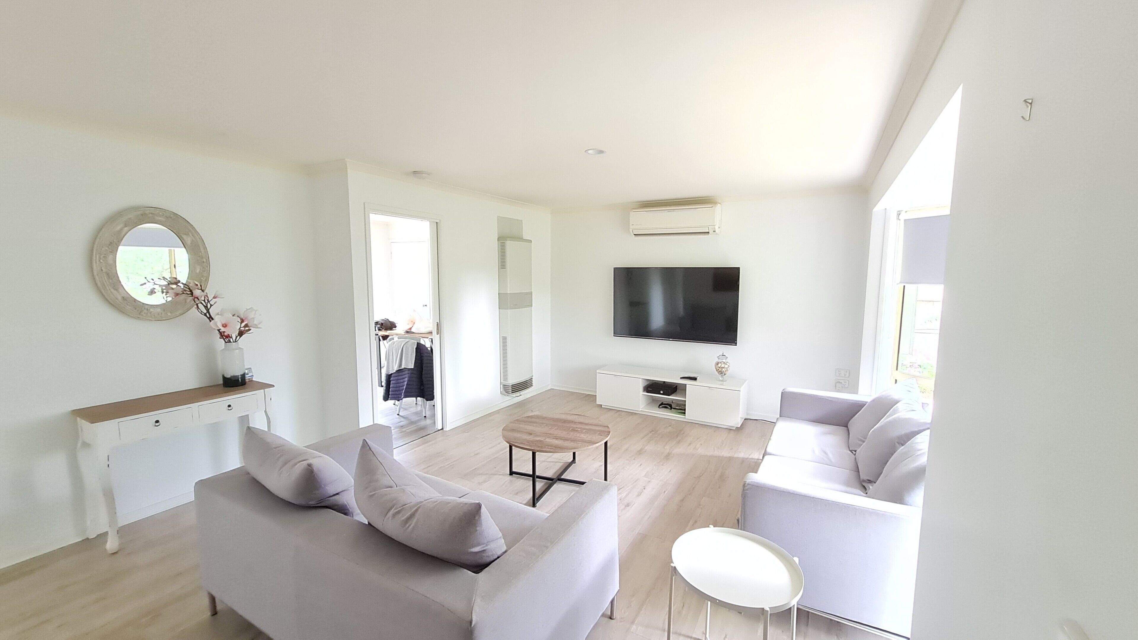 A modern living room with a grey sofa, a white armchair, and a coffee table. A flat-screen TV is mounted on the wall, and there is a white cabinet below it.