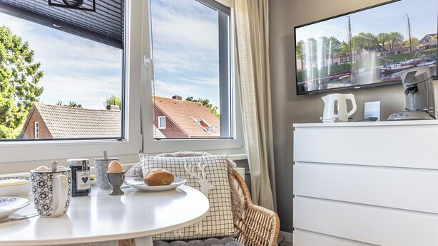 Quarto com mesa de jantar, cadeira de vime, penteadeira branca e TV exibindo uma paisagem.