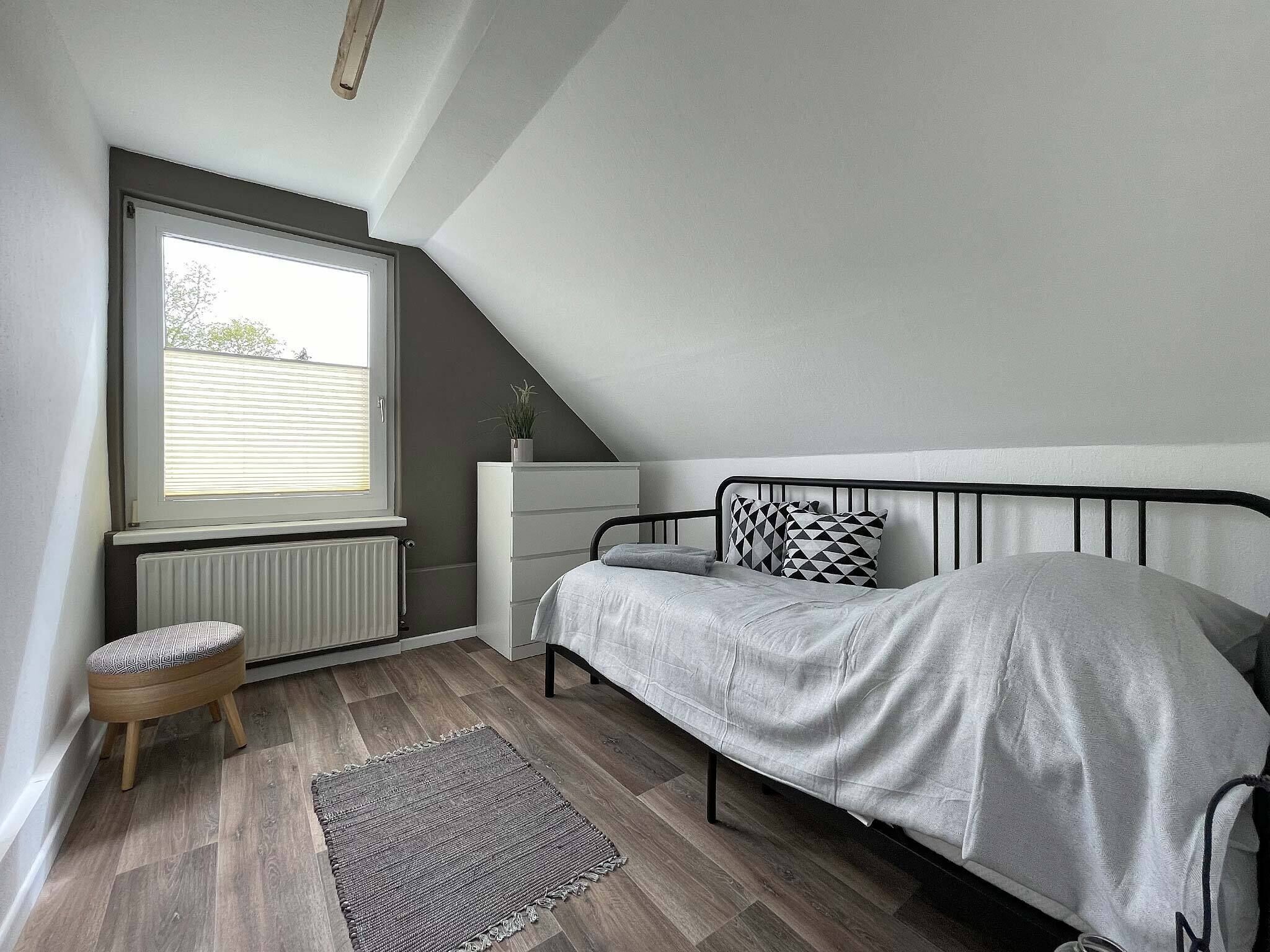 Makuuhuoneessa on sänky, lipasto, patteri, ikkuna ja pieni jakkara.