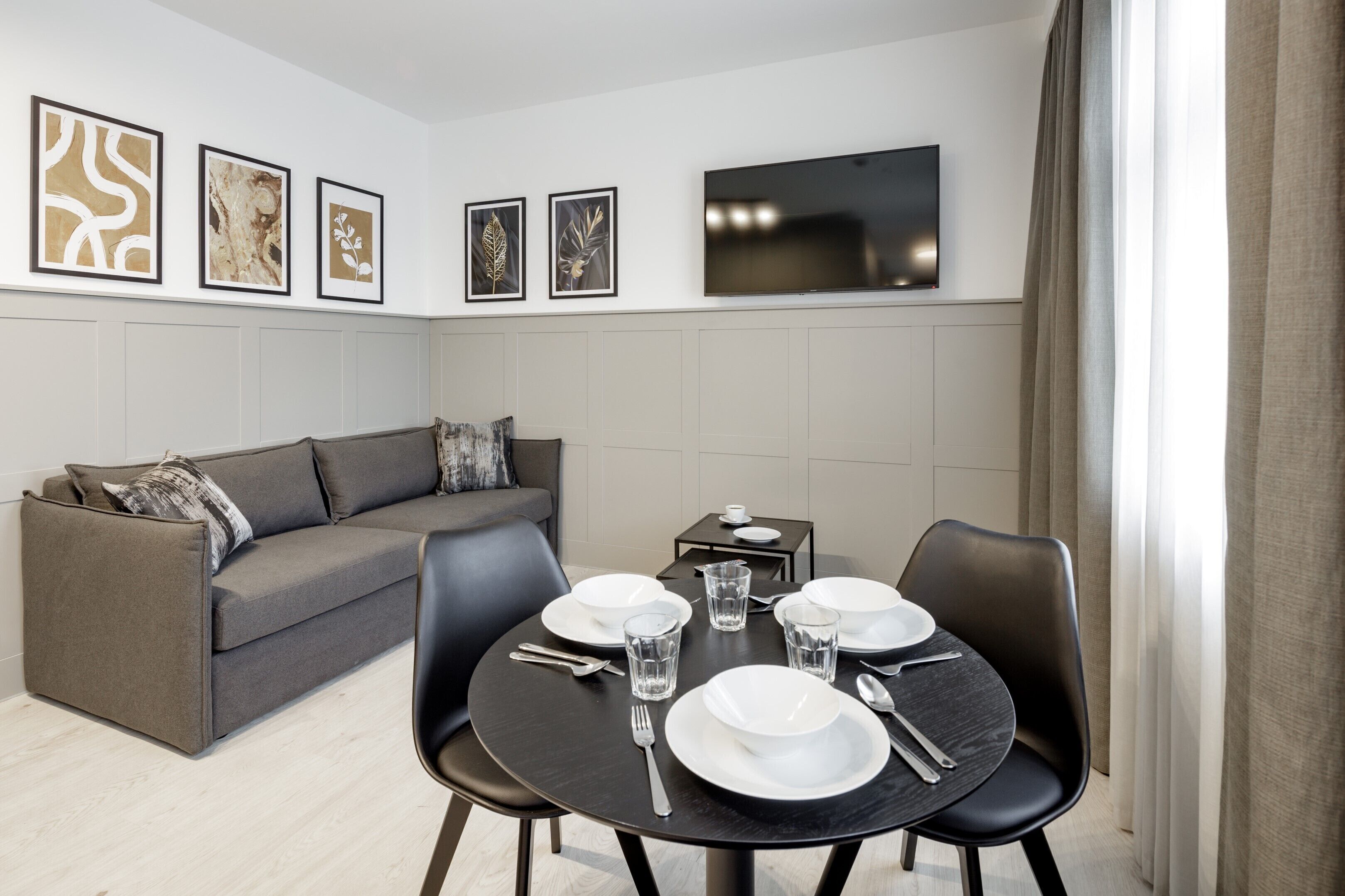 Ein modernes Wohnzimmer mit einem grauen Sofa, einem Esstisch mit weißen Tellern und Gläsern sowie einem an der Wand montierten Flachbildfernseher.