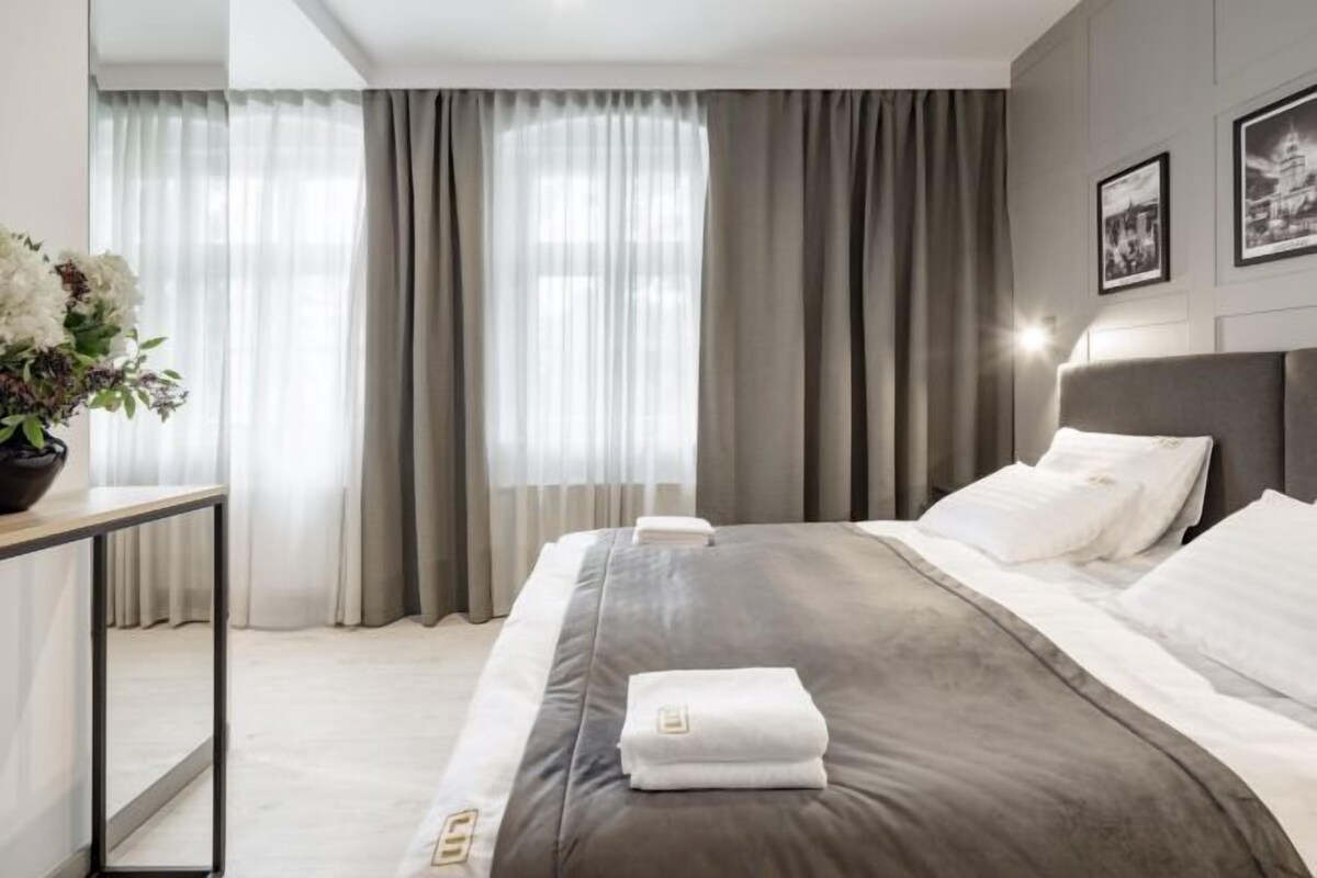 A hotel room with a bed, curtains, a bedside table with a vase, and a wall-mounted light.