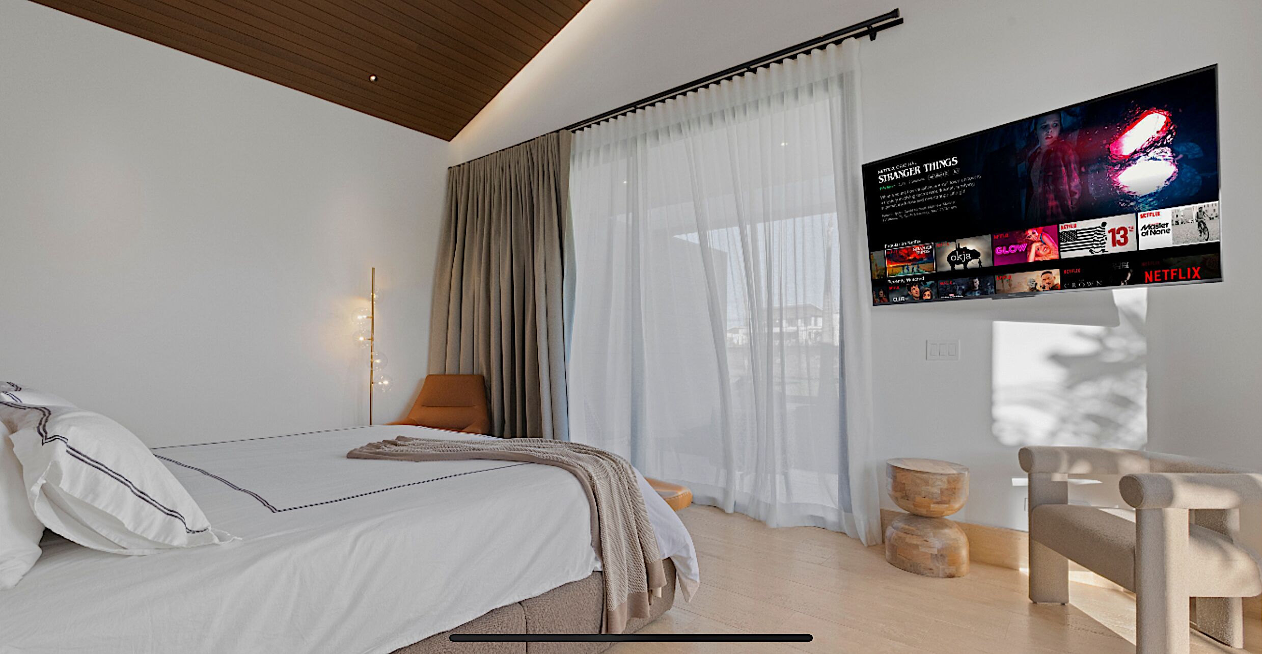 Una habitación de hotel moderna con una cama grande, un televisor de pantalla plana montado en la pared, techo de madera y un amplio ventanal con cortinas ligeras.