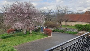 A paved driveway leading to a house with a brick roof, surrounded by a garden with blooming trees and shrubs. - Chambre avec cuisine équipée sur demande (Figeac)