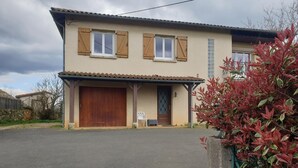 A two-story house with a garage, a covered porch, and a small garden with red-leaved plants. - Chambre avec cuisine équipée sur demande (Figeac)