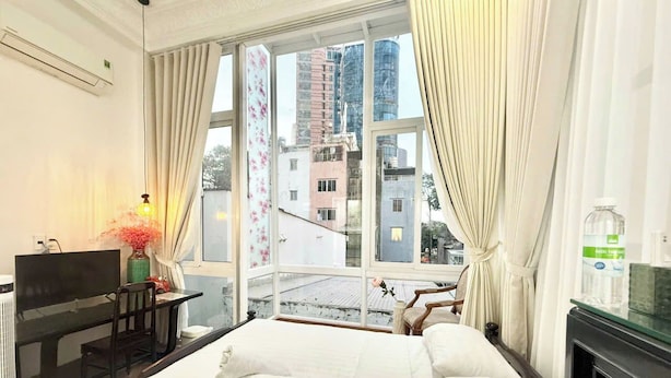 Quarto de hotel com cama, escrivaninha com televisão, janela com cortinas e vista para prédios.