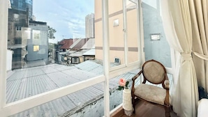 Quarto com uma grande janela com vista para a paisagem urbana, uma cadeira de madeira e uma mesa pequena com um vaso.