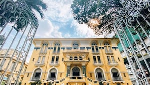 Um edifício amarelo grande com entrada central, varandas e uma fonte em frente.