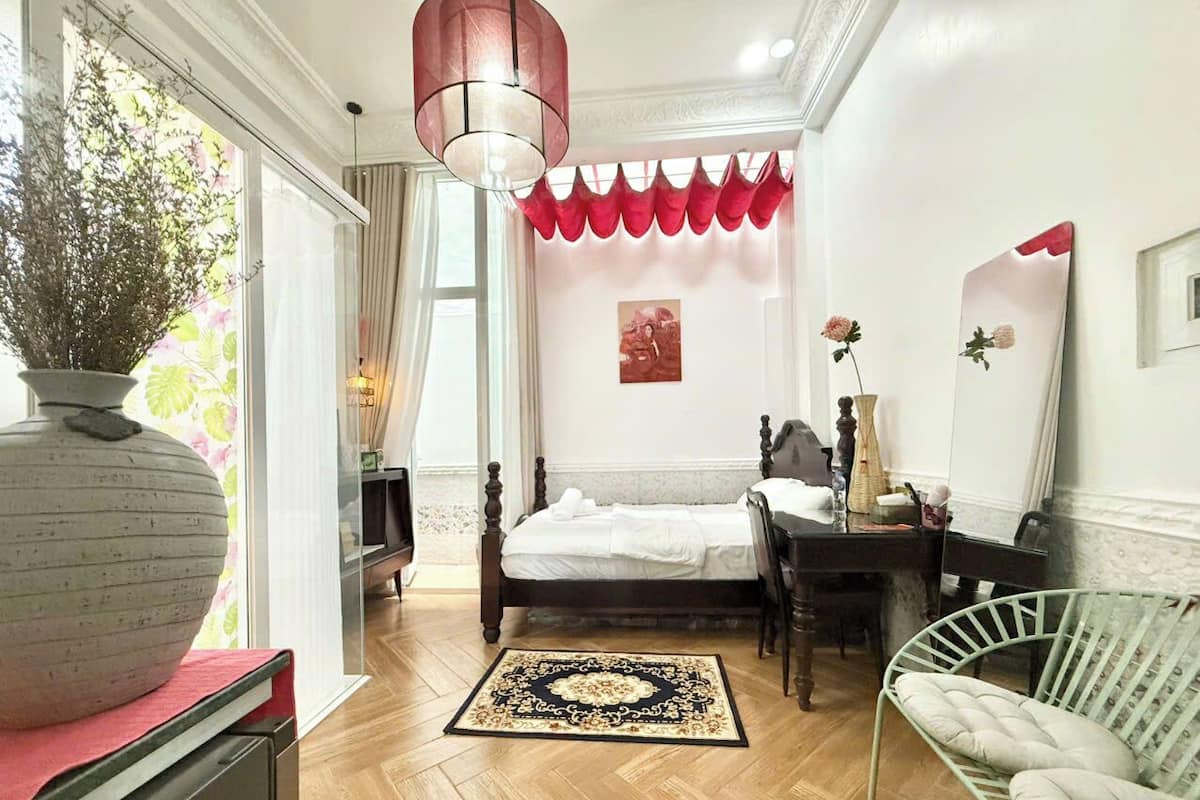 Un dormitorio con piso de madera, una cama con ropa de cama blanca, una mesita de noche, una silla, un florero grande y una ventana con cortinas.