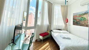 Quarto de hotel com uma cama, escrivaninha, cadeira e janela com cortinas.