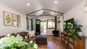 A cozy living room with a brown sofa, a wooden coffee table, and a TV on a wooden stand. There is a green door leading outside, and a fireplace with a black chimney. - Upon a Star - Welcoming you to the most charming holiday retreat, nestled in the picturesque village of Wandiligong, Victoria. (Wandiligong)