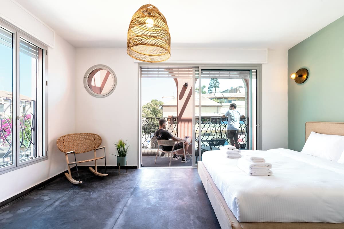 A modern hotel room with a large bed, a rocking chair, a potted plant, and a view of the outside balcony.