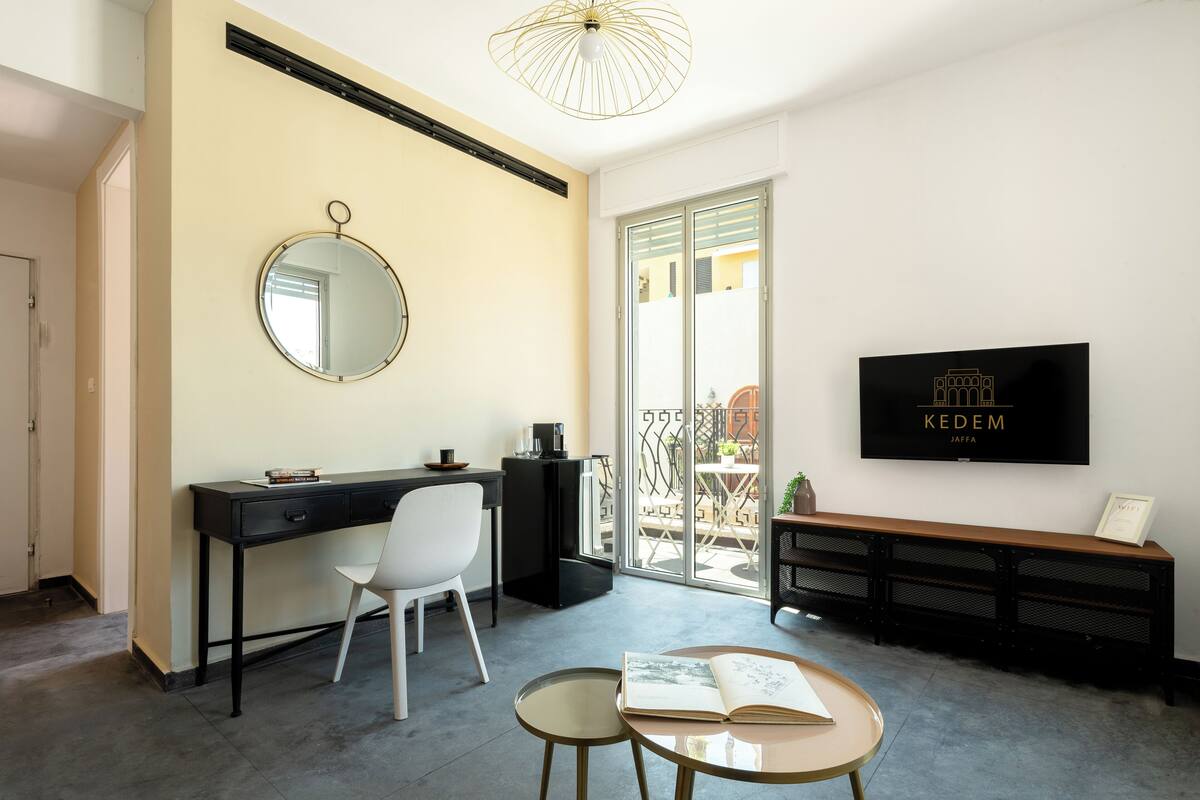 A modern living room with a black desk, white chair, and a round table with an open book. A TV is mounted on the wall, and a bench is against the wall.