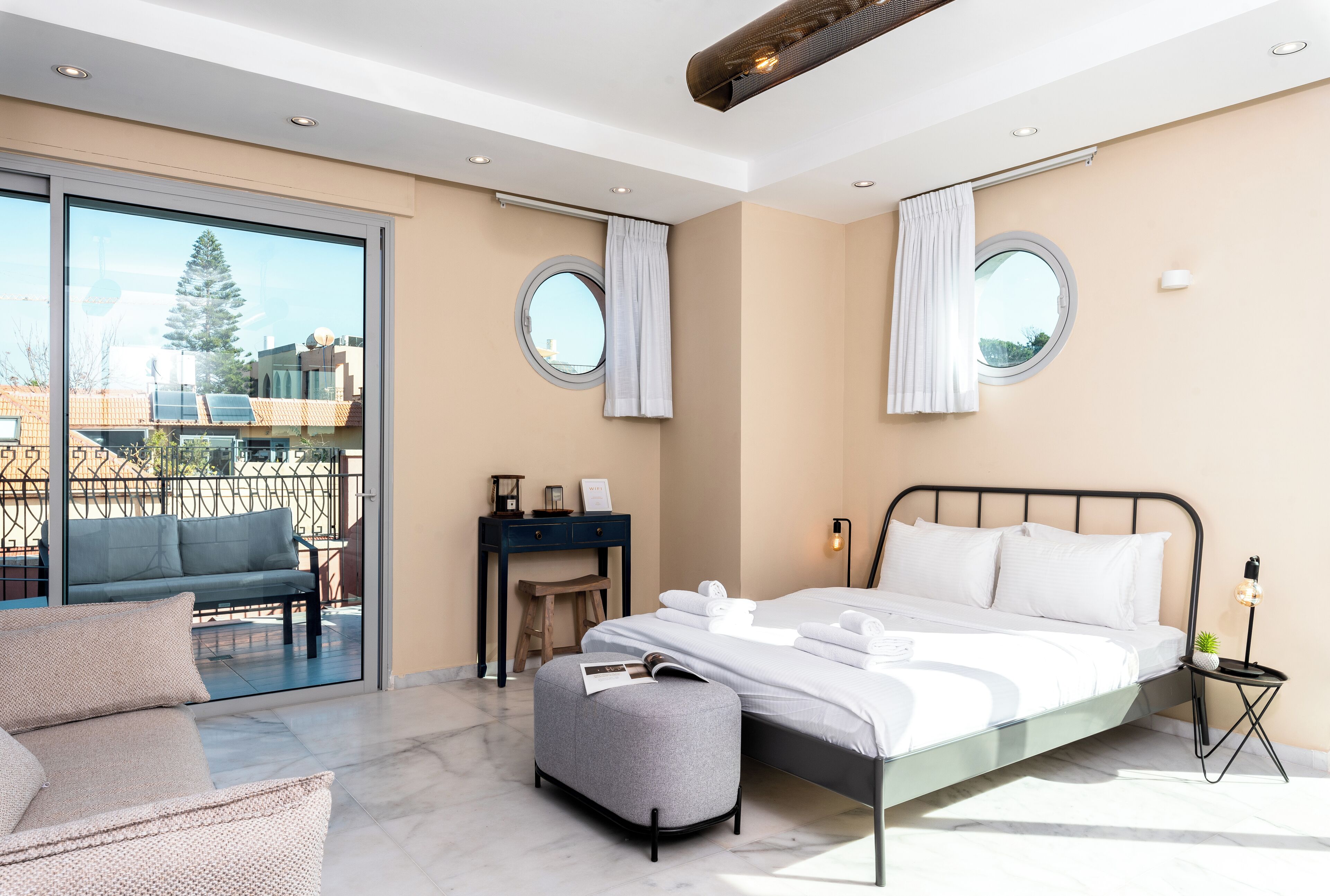 A modern bedroom with a bed, a desk, a chair, and a balcony with a view of buildings and a tree.