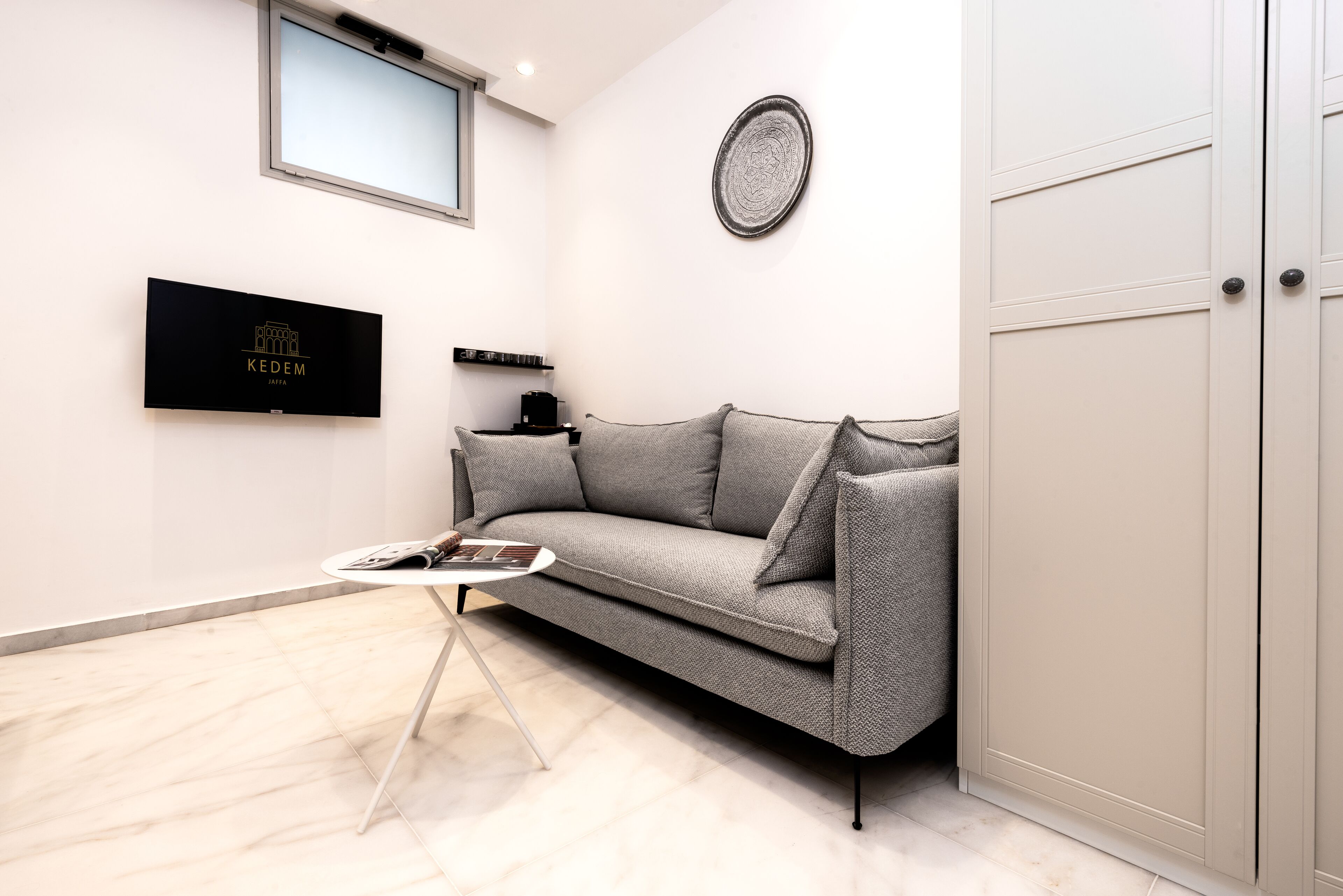 A modern living room with a grey sofa, a small round coffee table, a flat-screen TV mounted on the wall, and a cabinet with a door.
