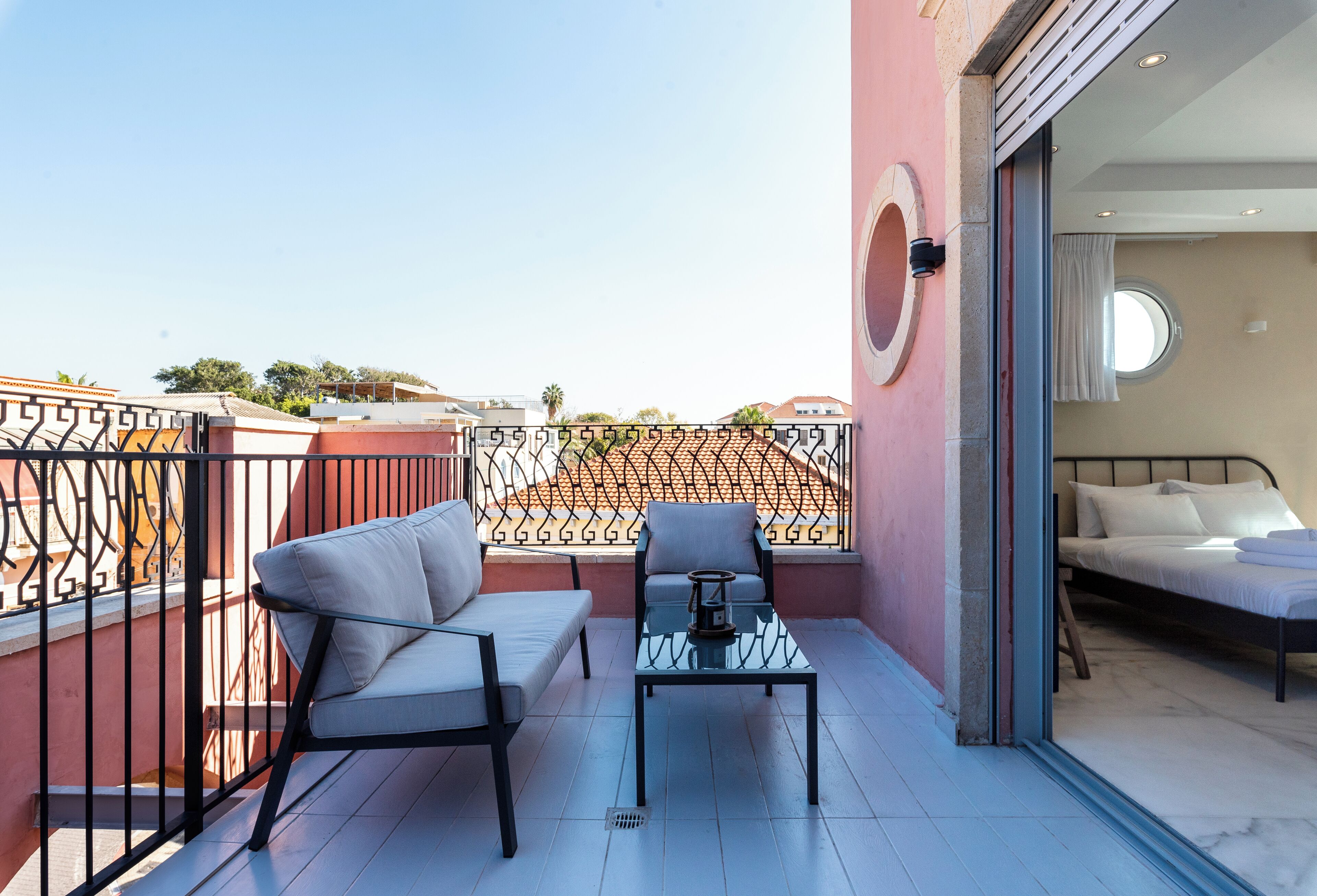 A balcony with a sofa and a table, a bed inside the room, and a view of the cityscape.