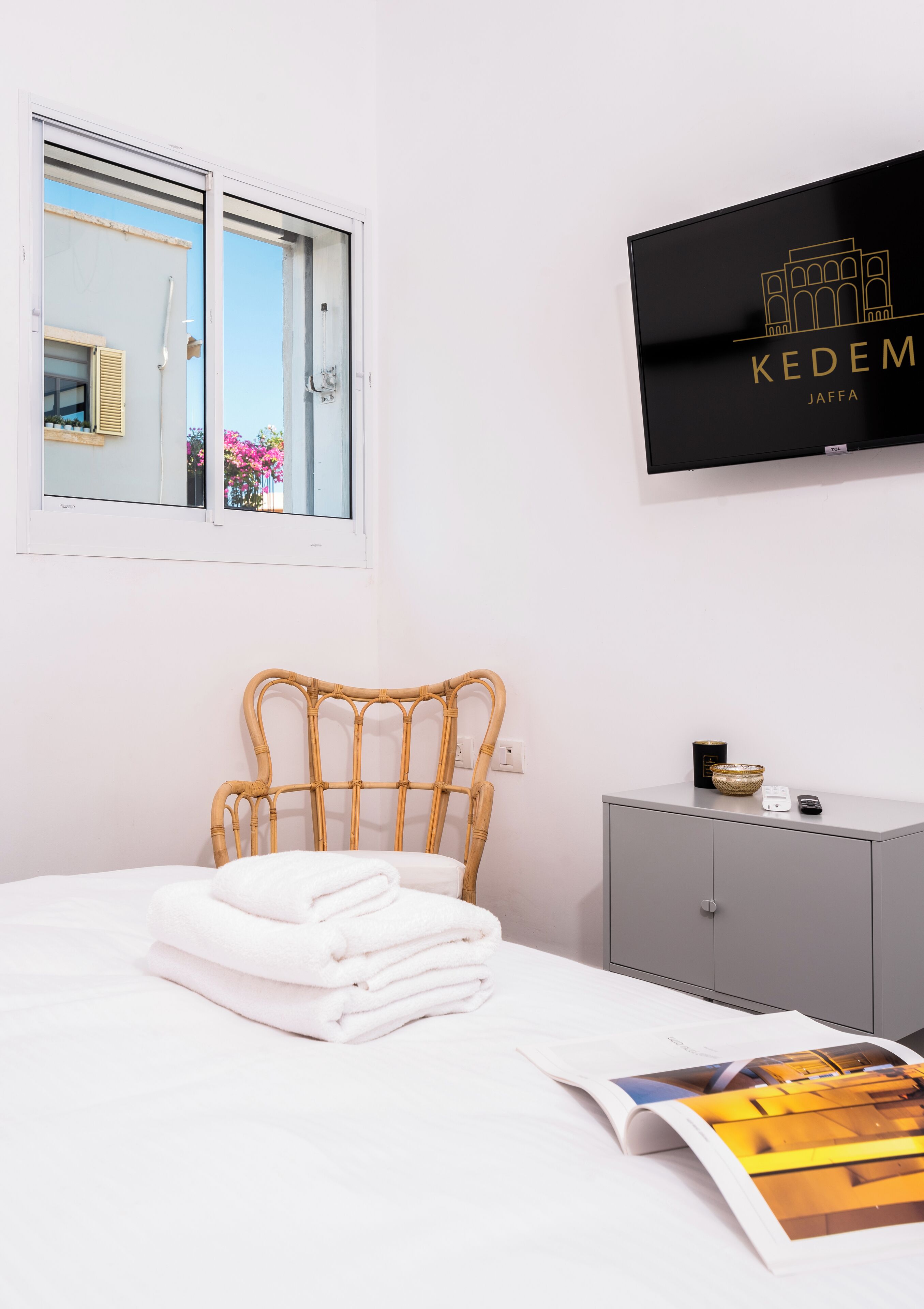 Una cama bien hecha con toallas blancas dobladas, un mueble gris con un cuenco y un control remoto, y un cartel enmarcado en la pared.
