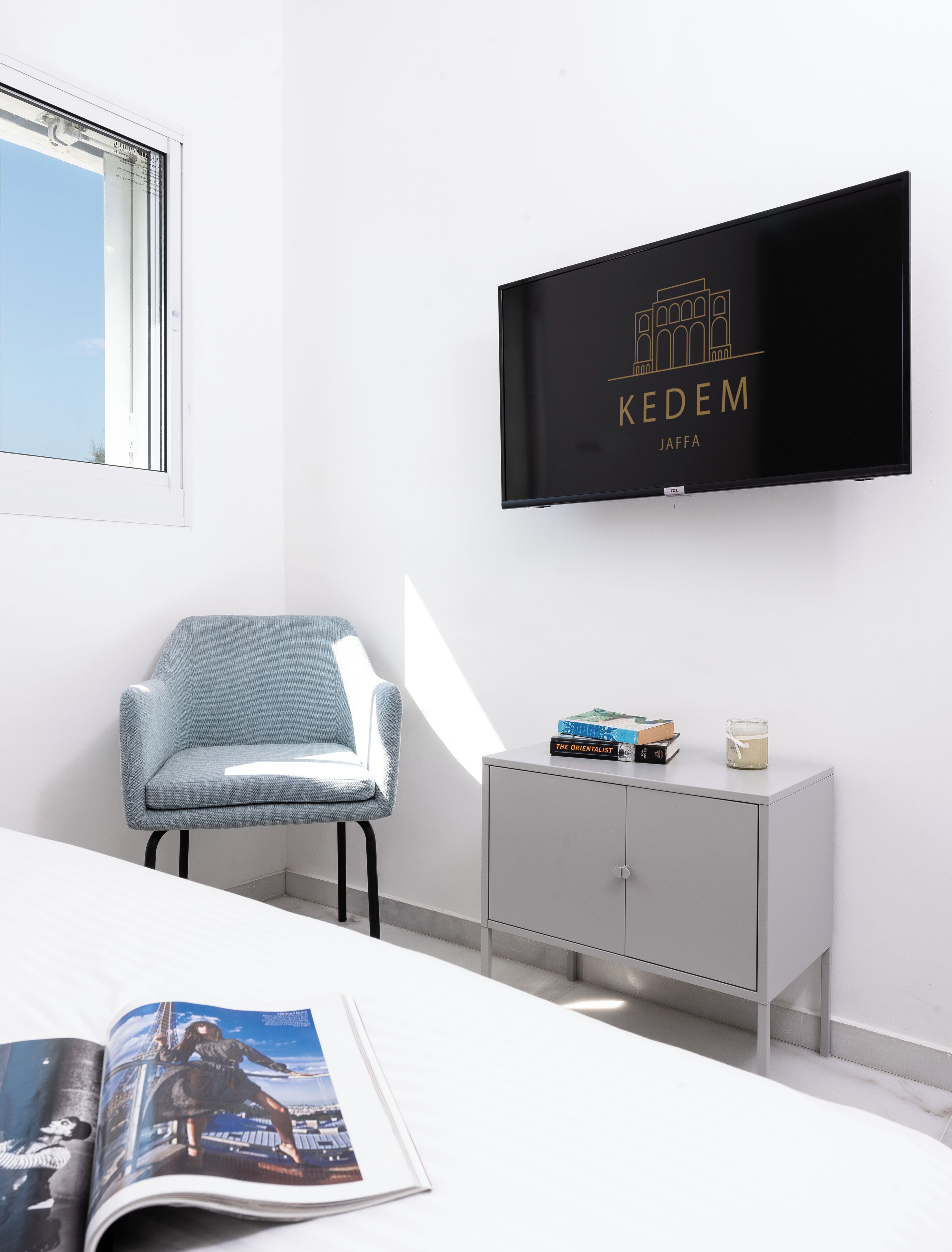 A modern room with a TV displaying 'KEDEM JAFFA', a grey chair, a small cabinet with books and a candle, and a magazine on the table.