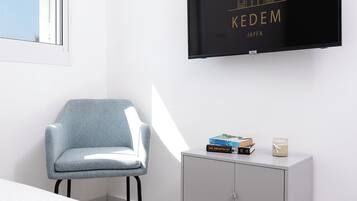 A modern room with a TV displaying 'KEDEM JAFFA', a grey chair, a small cabinet with books and a candle, and a magazine on the table.