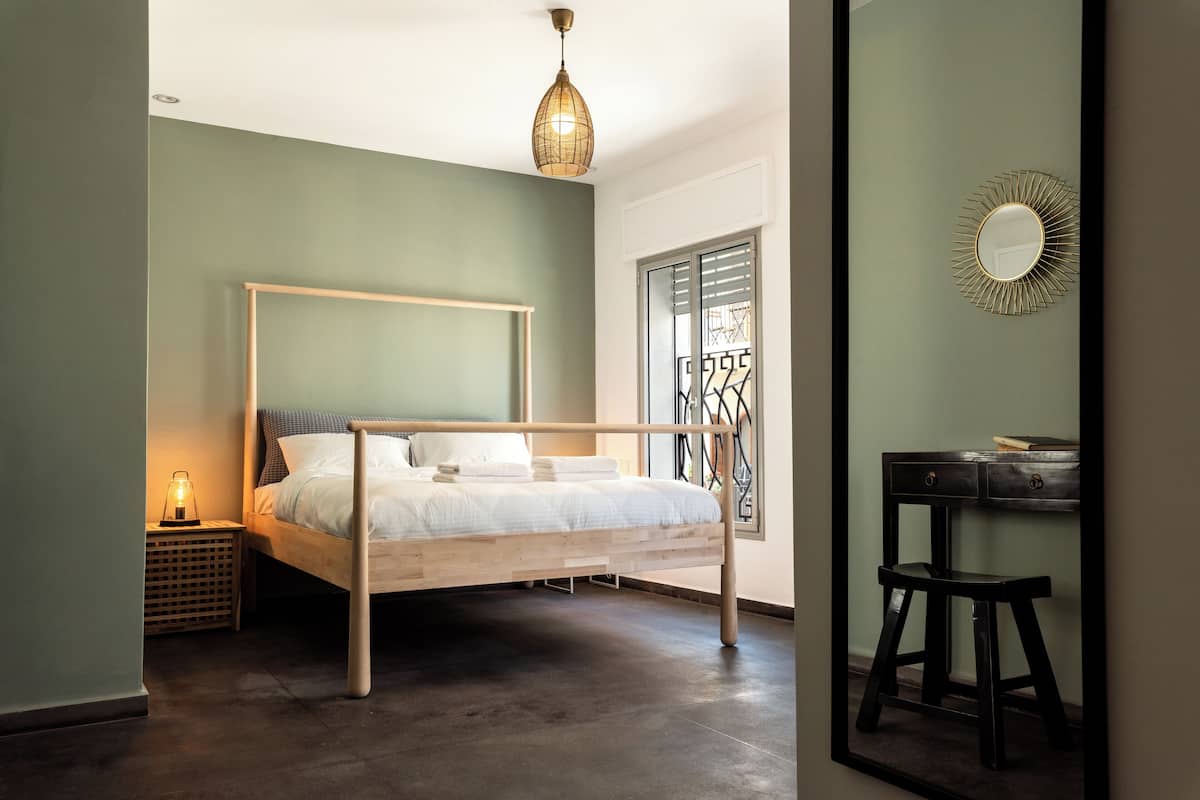 A bedroom with a wooden bed, a nightstand with a lamp, a desk with a chair, and a window with blinds.