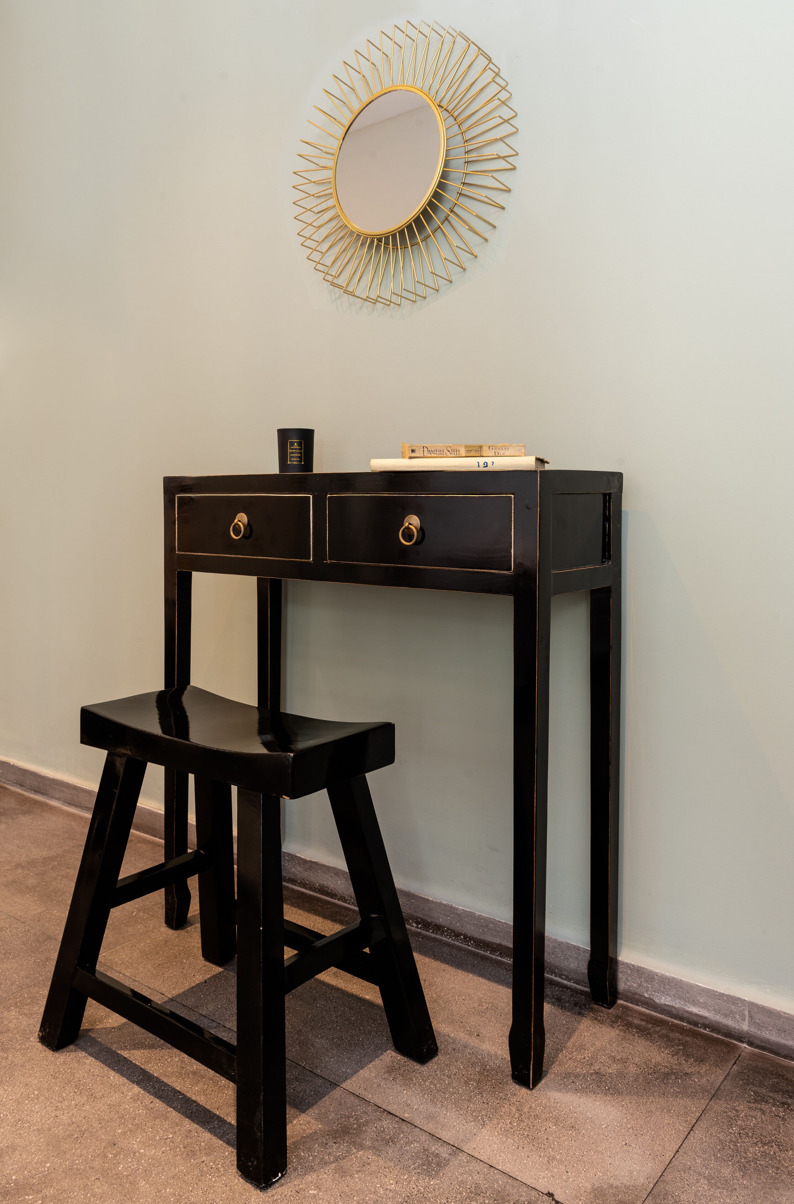 Un bureau noir avec deux tiroirs et un tabouret, un miroir encadré d’or accroché au mur, et une petite table sur laquelle se trouvent des livres et une tasse.