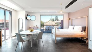 Une chambre moderne avec un grand lit, une table à manger avec des chaises et un balcon offrant une vue sur les immeubles.