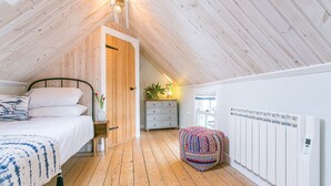 Una camera da letto mansardata accogliente, con soffitto spiovente, letto con biancheria bianca, una cassettiera, un poggiapiedi colorato e un termosifone bianco.